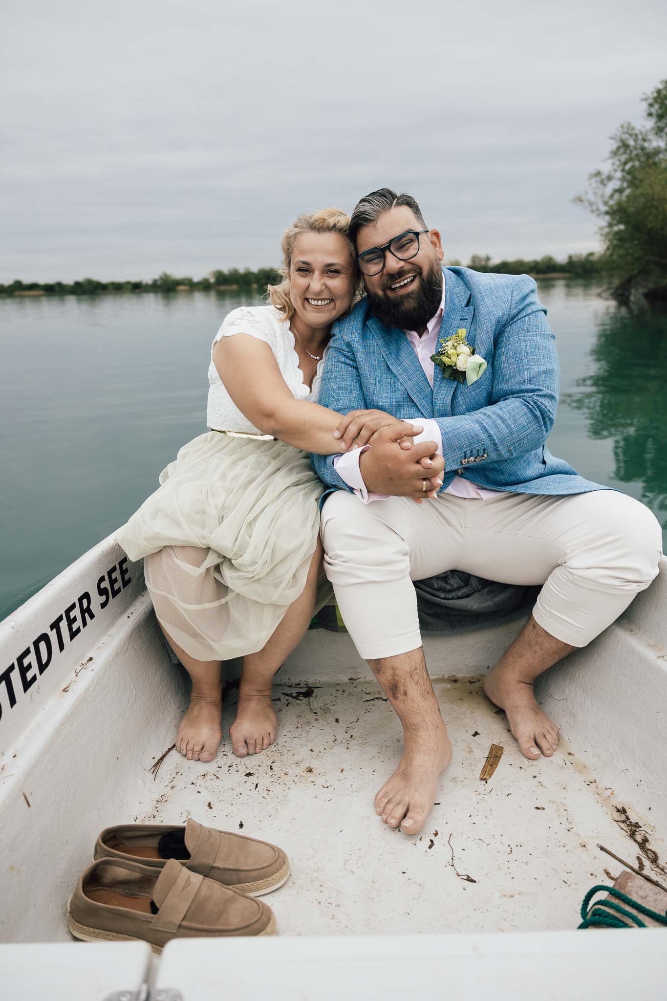 Barfüssiges Brautpaar sitzt lachend in einem Ruderboot auf dem See
